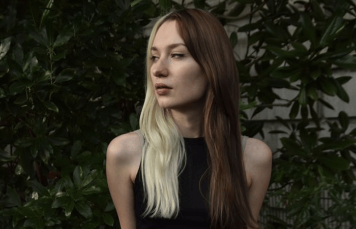A girl with half brown and half blonde hair, standing in front of a wall of leaves, wearing a black top. She looks to her right, with a serious expression.